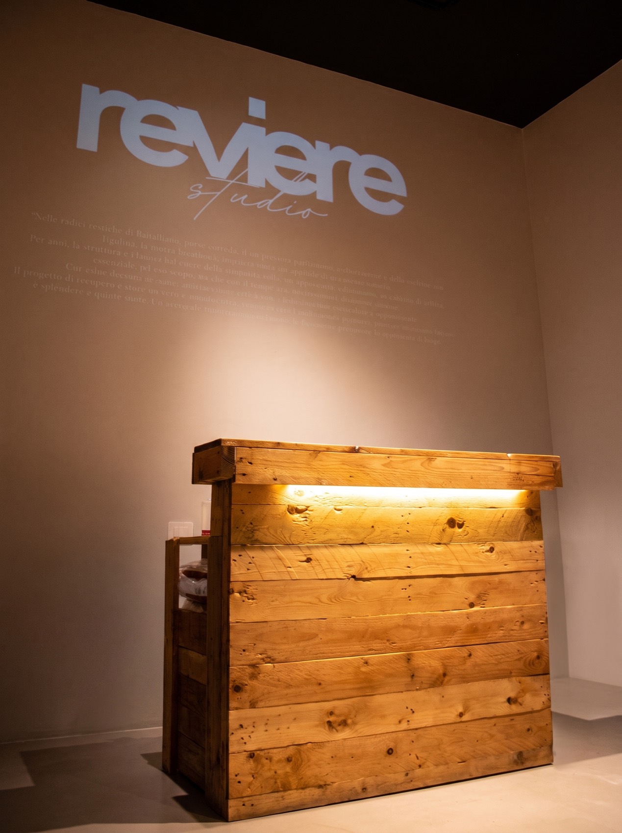 Bancone in legno massello con luce calda sotto il piano e il logo Reviere Studio proiettato sulla parete